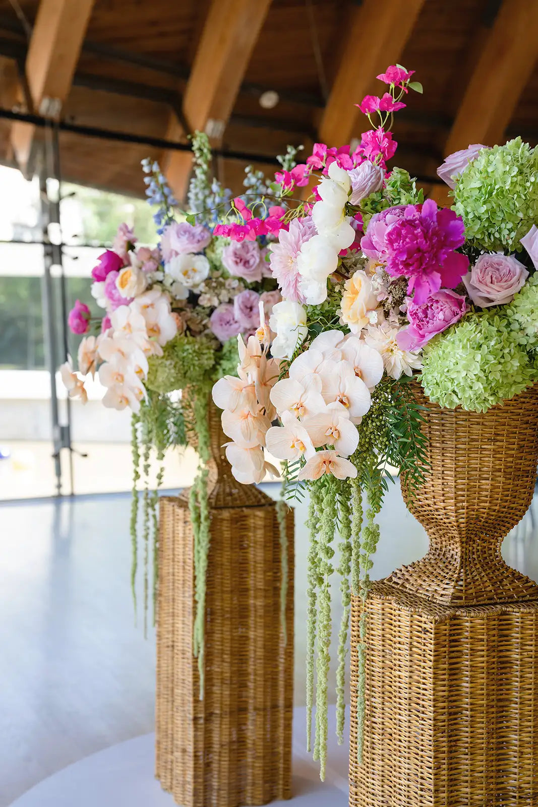 Decorative floral arrangements in rattan urns on stands with a blurred indoor background.