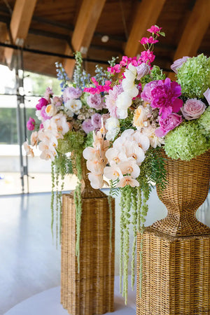 Decorative floral arrangements in rattan urns on stands with a blurred indoor background.