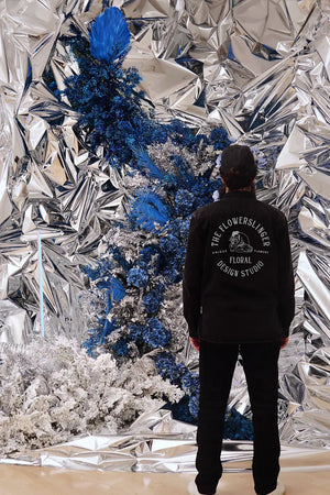 Person wearing a black Longsleeve Shirt with text standing in front of a reflective, silver, and blue floral installation.