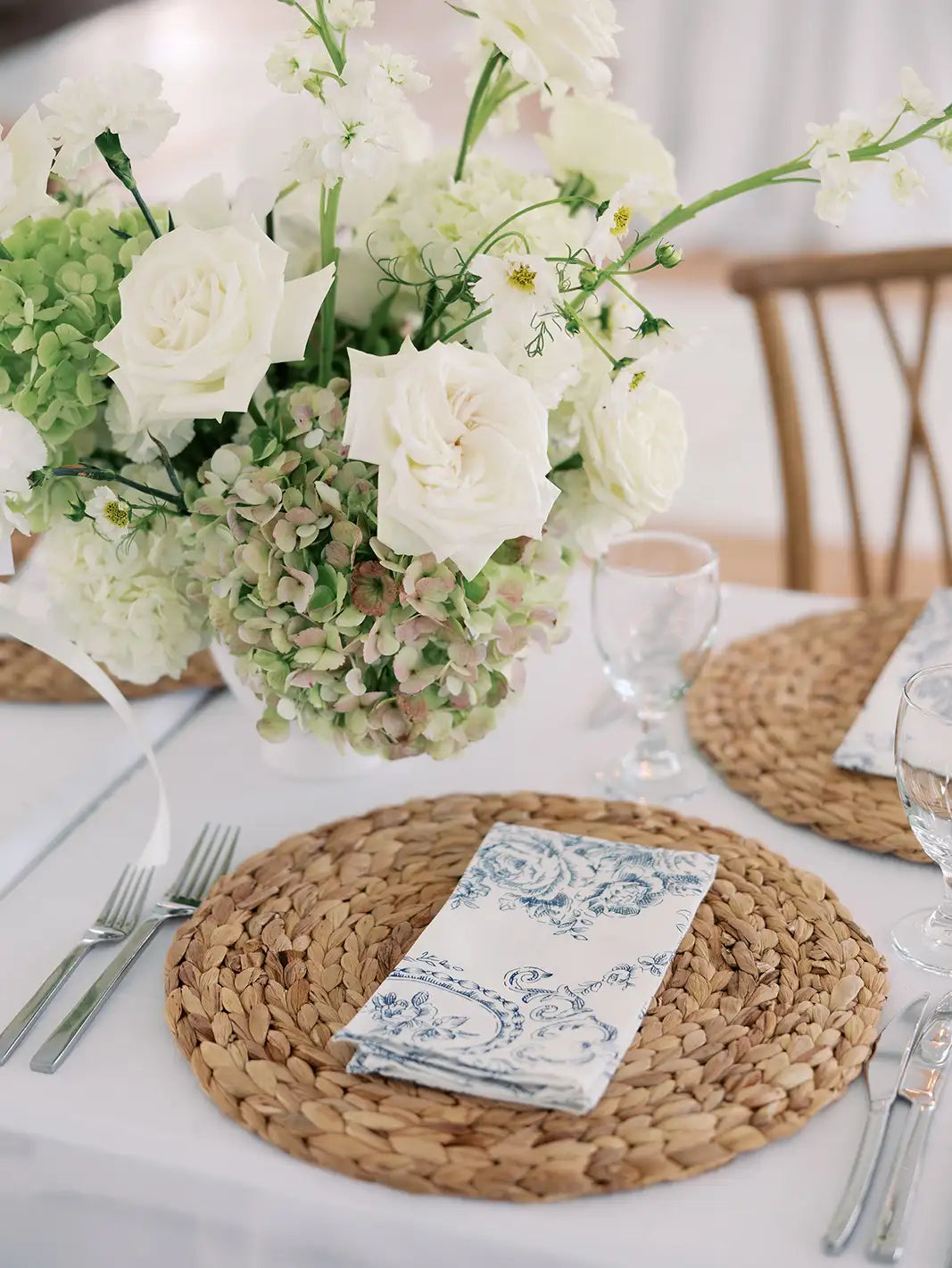 Table setting with woven placemats, floral centerpiece, and cutlery on a white tablecloth.