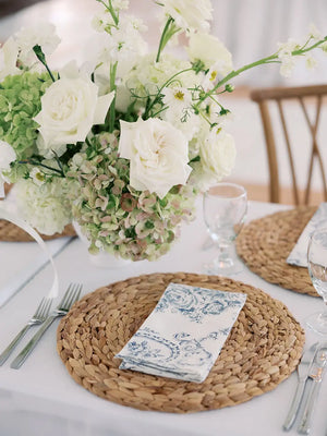 Table setting with woven placemats, floral centerpiece, and cutlery on a white tablecloth.