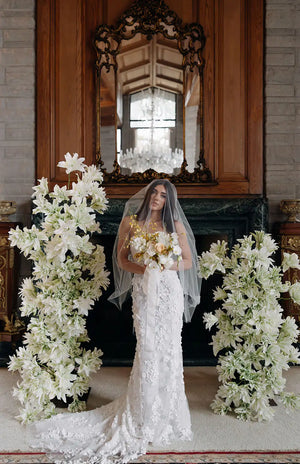Editorial Bride in a white wedding dress holding flowers in a decorated room with floral arrangements and a mirror.