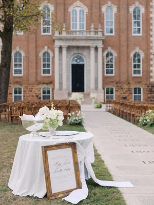 Decorative table setup with flowers and a framed sign in front of a brick building.