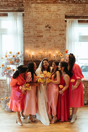 Bride with bridesmaids in pink dresses in a room with brick wall and candles holding flowers with ceremony flowers behind them.