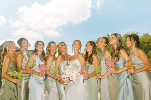 Group of women in light-colored dresses holding flowers against a blue sky.