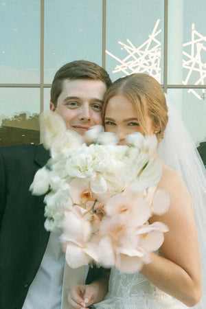 Couple holding a bouquet of flowers in front of a light blue window with decorative lights.