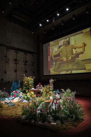 Diorama setup with floral arrangements and a screen displaying a person.