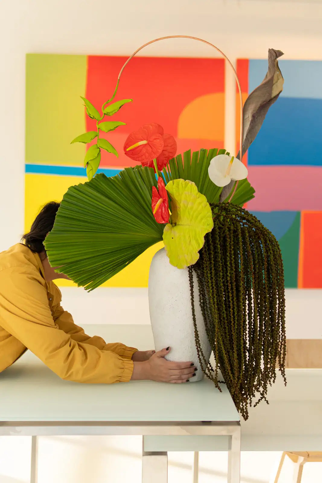 Person arranging a floral arrangement in a white vase against a colorful abstract painting.